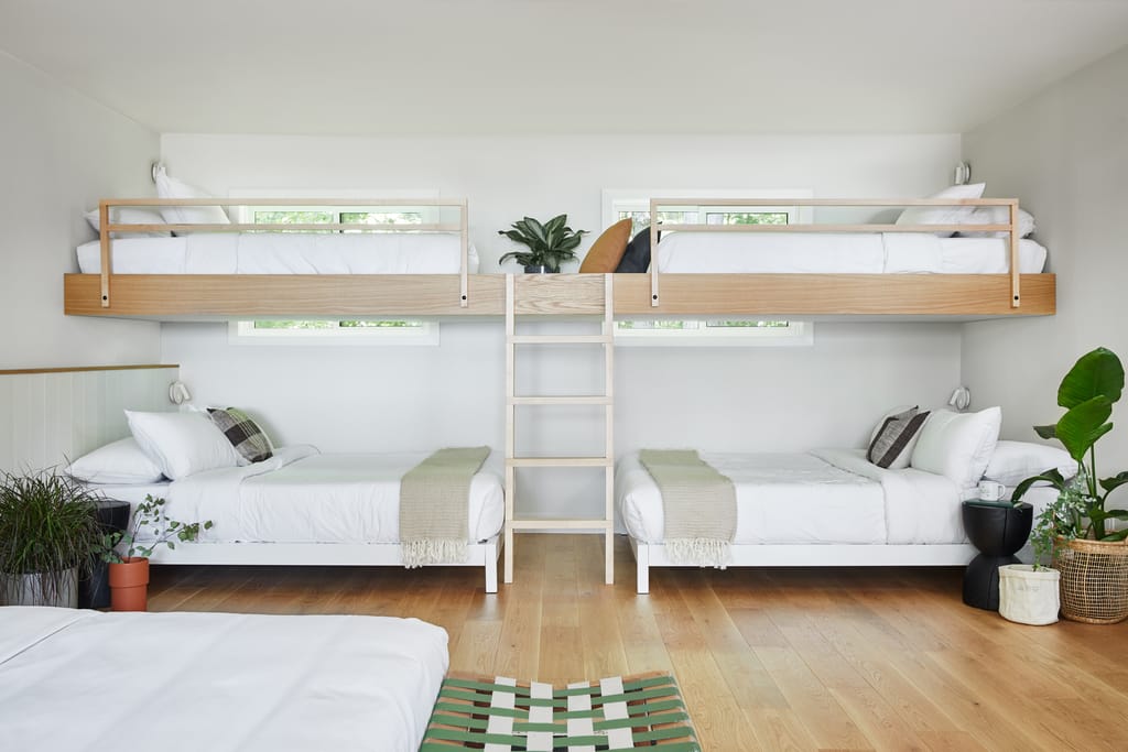 Room with two built-in bunk beds above a pair of beds, plants and a woven bench