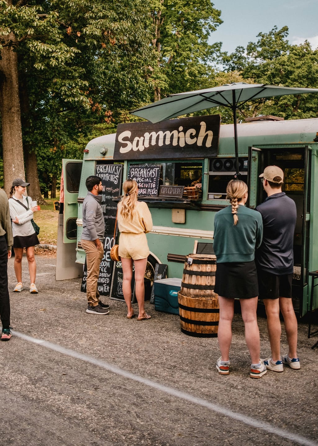 People lined up at a mint-green food truck with a Sammich sign under the trees
