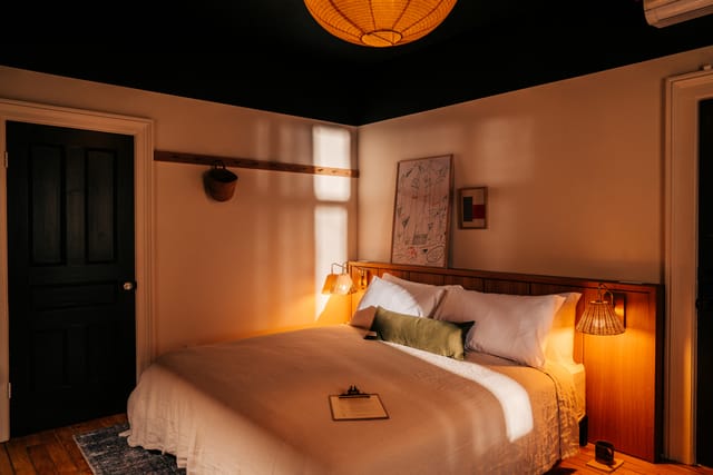 Guest room with a low bed, rattan pendant, framed art and a dark ceiling lit by warm afternoon sun