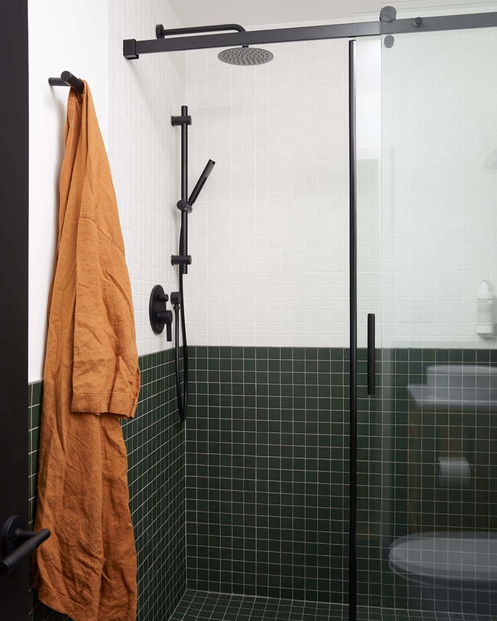 Walk-in shower with green and white tile, black fixtures and a rust linen robe on a hook
