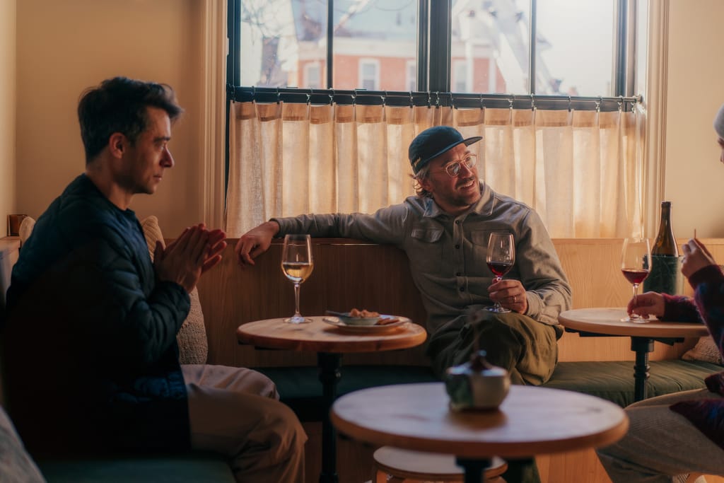 Two men talk over glasses of wine at small round tables on a banquette by a curtained window