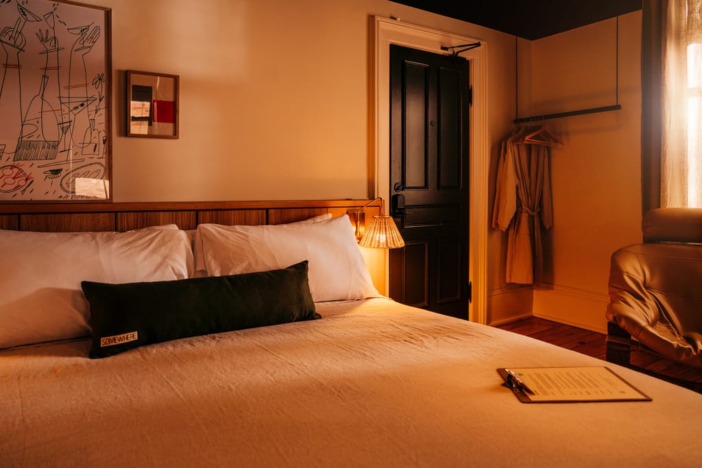 Bed with a dark bolster pillow, framed art, a robe on a rail and a clipboard menu on the duvet