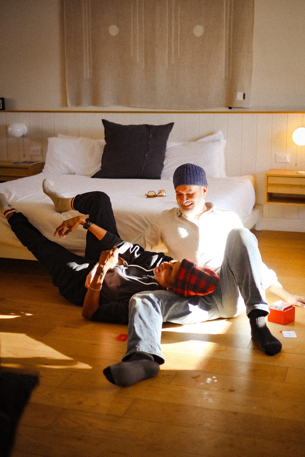 Two guests laughing on the sunlit floor of a guest room, leaning against the bed