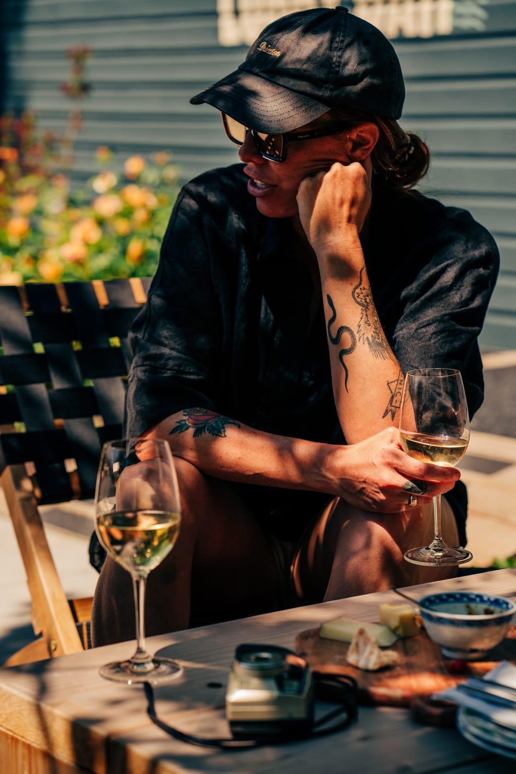 Person in a cap and sunglasses holding a glass of white wine at a patio table with a cheese board