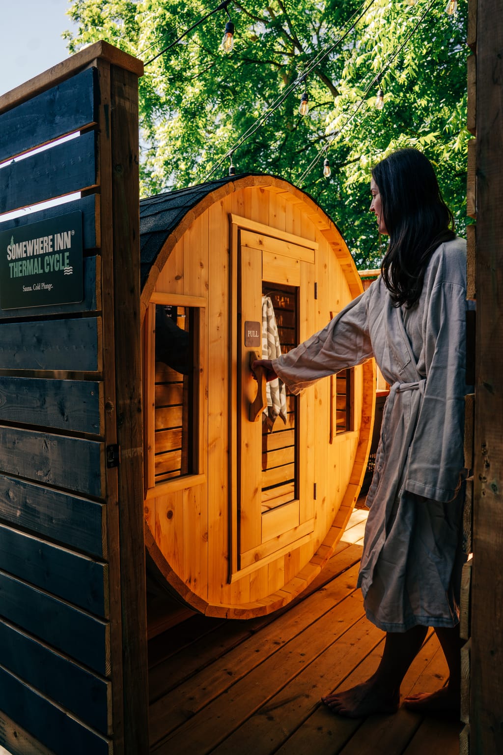 Woman in a robe opening the door of a barrel sauna beside a Thermal Cycle sign
