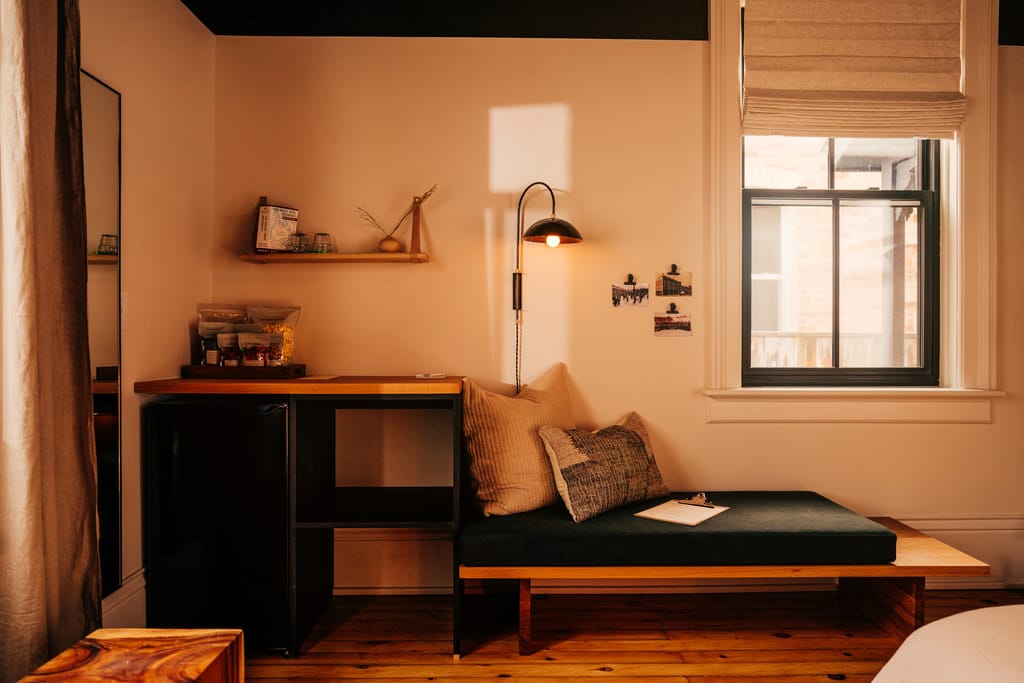 Corner of a guest room with a cushioned bench, desk, shelf, floor lamp and window