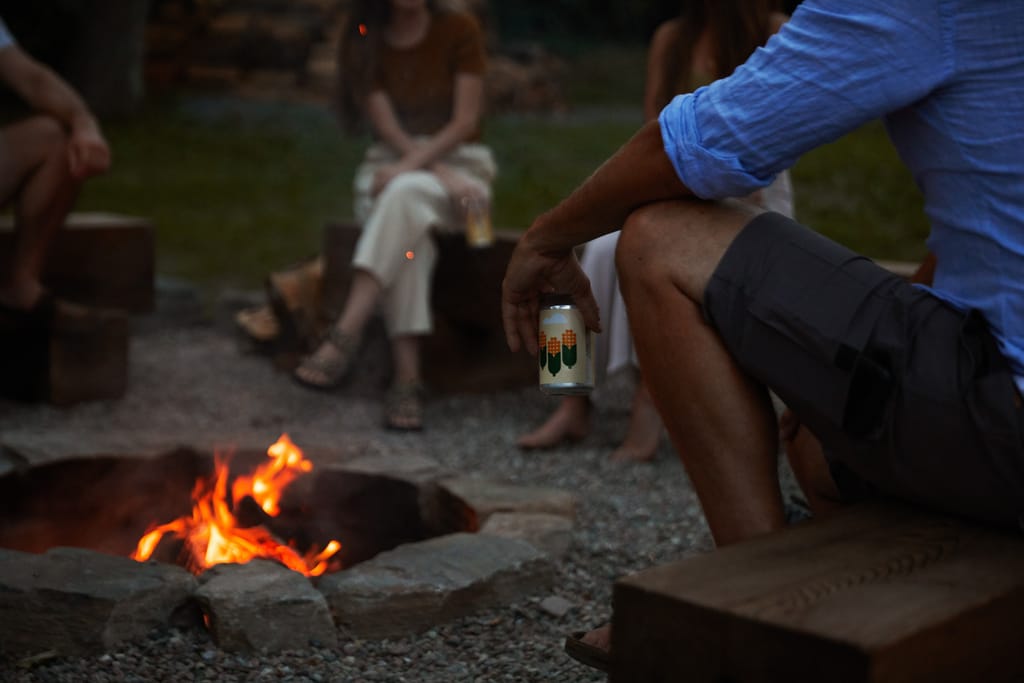 Fire in a stone-ringed pit, with a guest holding a can of beer on a timber bench