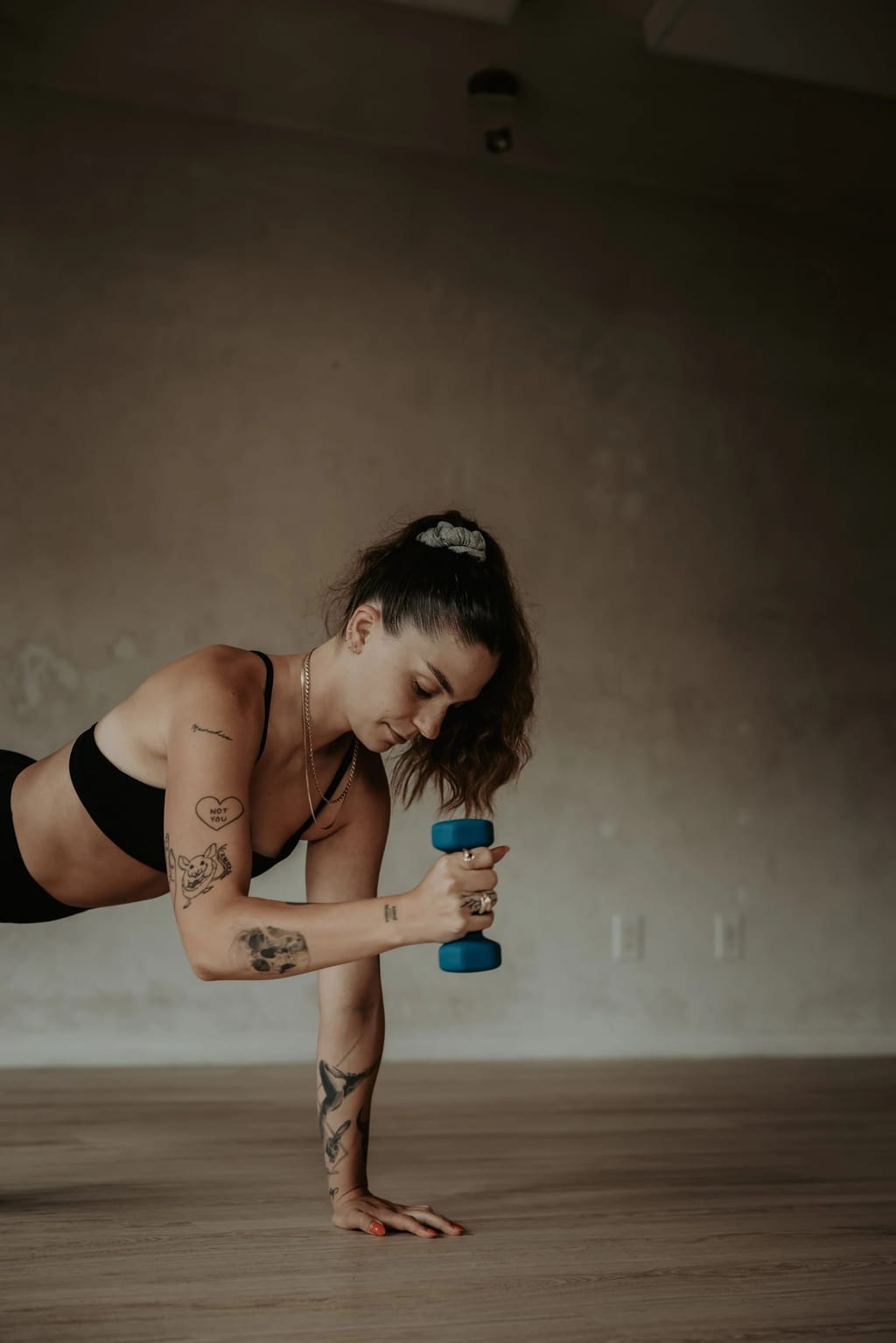 Woman in a plank holding a blue dumbbell in a bare-walled movement studio with a wood floor