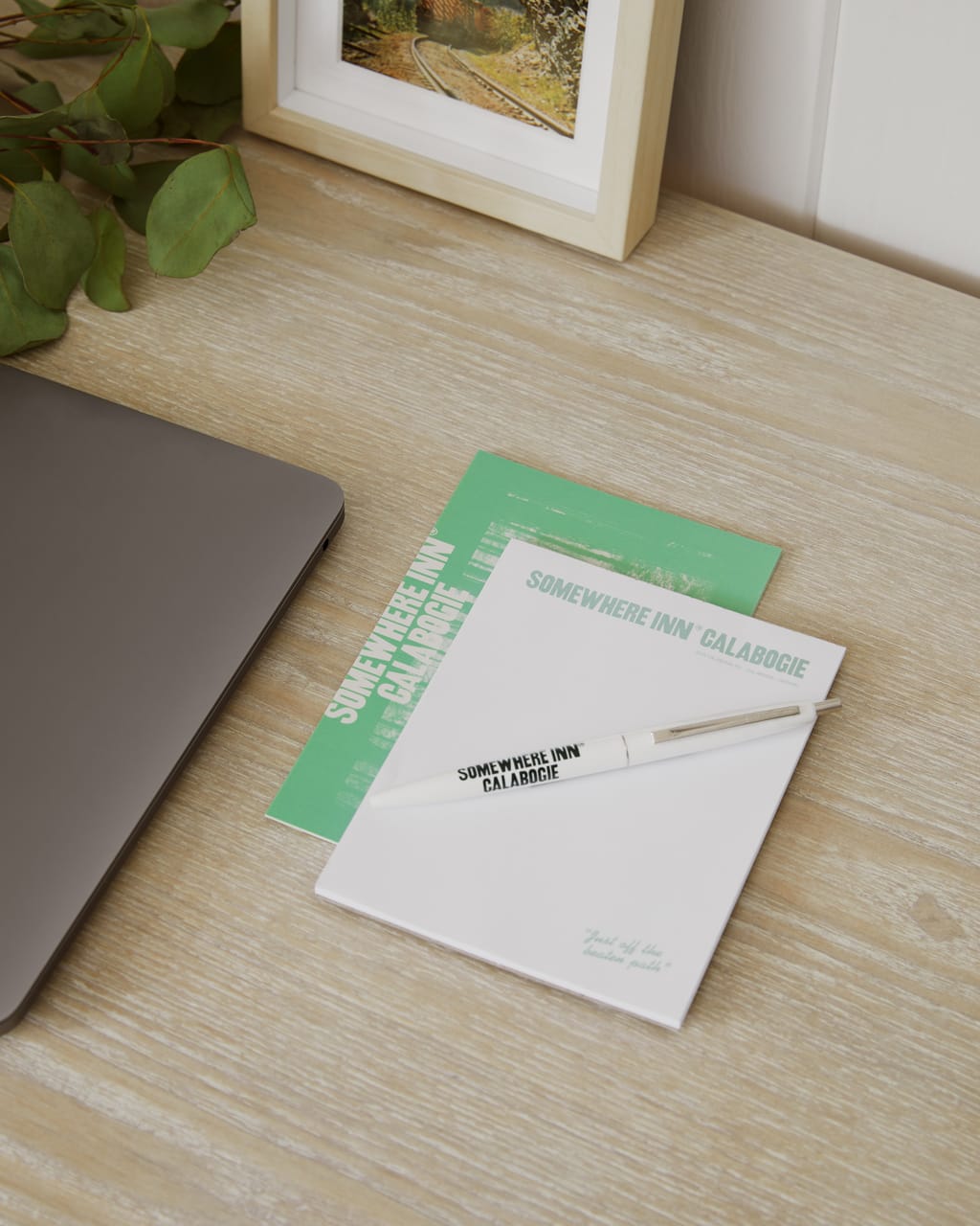 Somewhere Inn Calabogie notepads and a branded pen on a wood desk beside a laptop