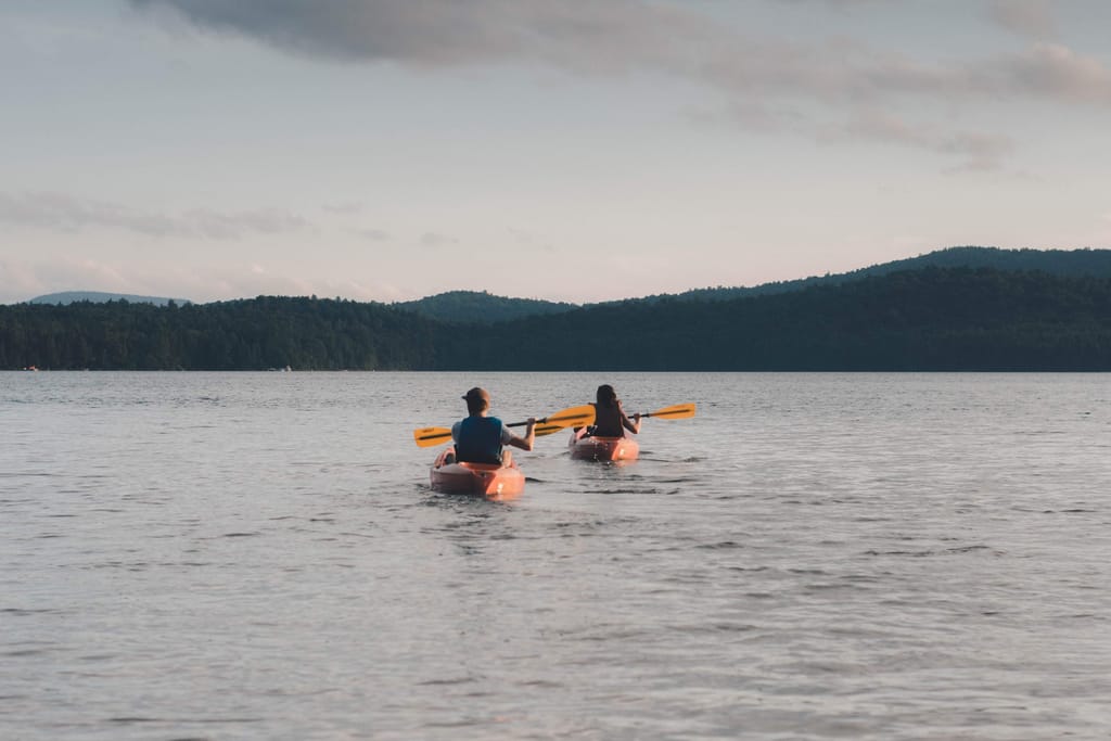 Two people paddling orange kayaks across a calm lake toward forested hills at dusk