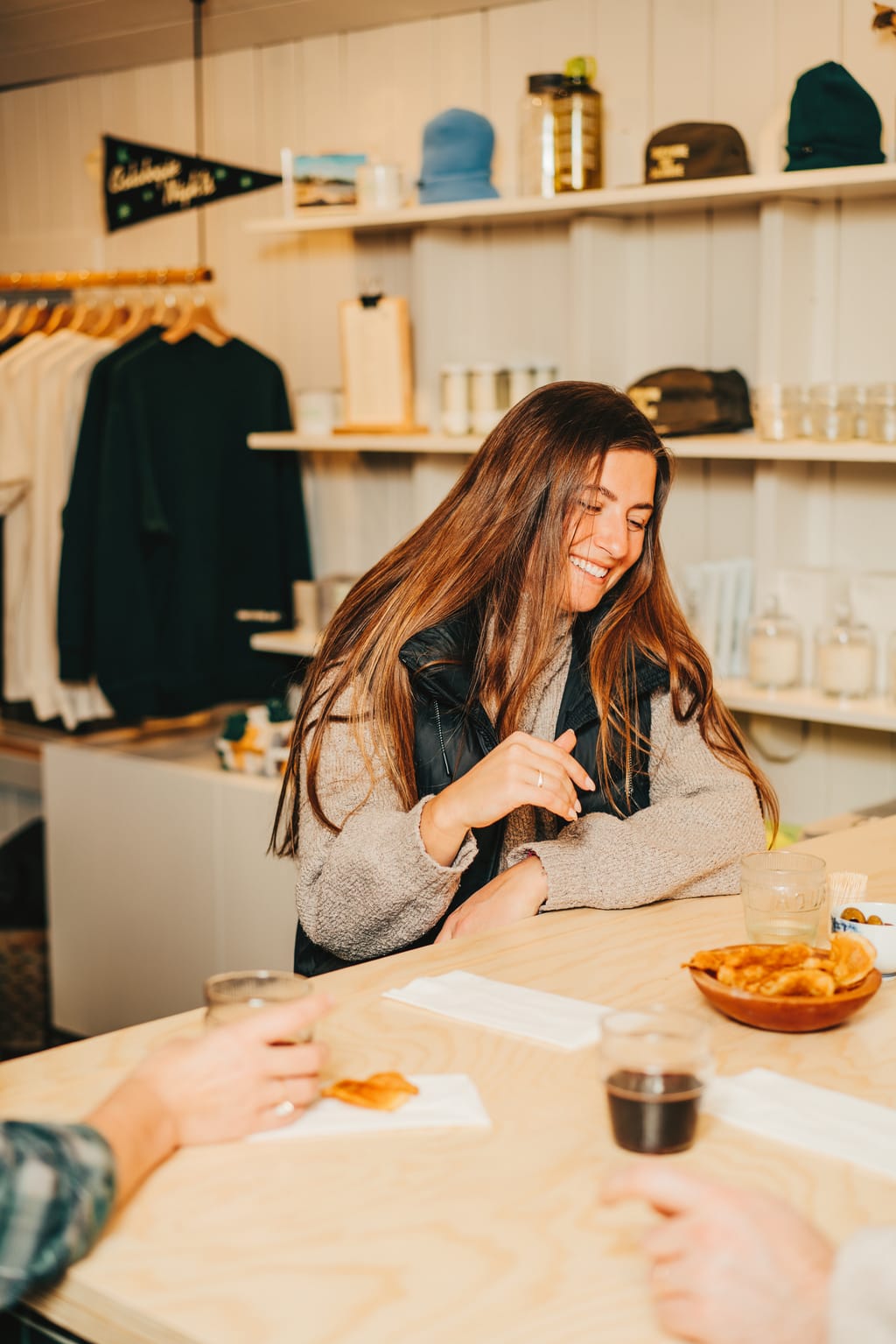 Smiling woman at a pale wood counter with snacks and drinks, a shop shelf with caps behind her