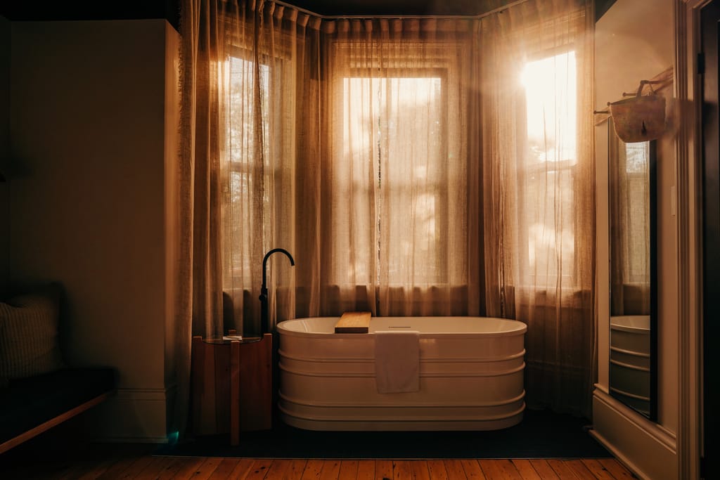Freestanding white tub in front of a sunlit bay window with sheer linen curtains