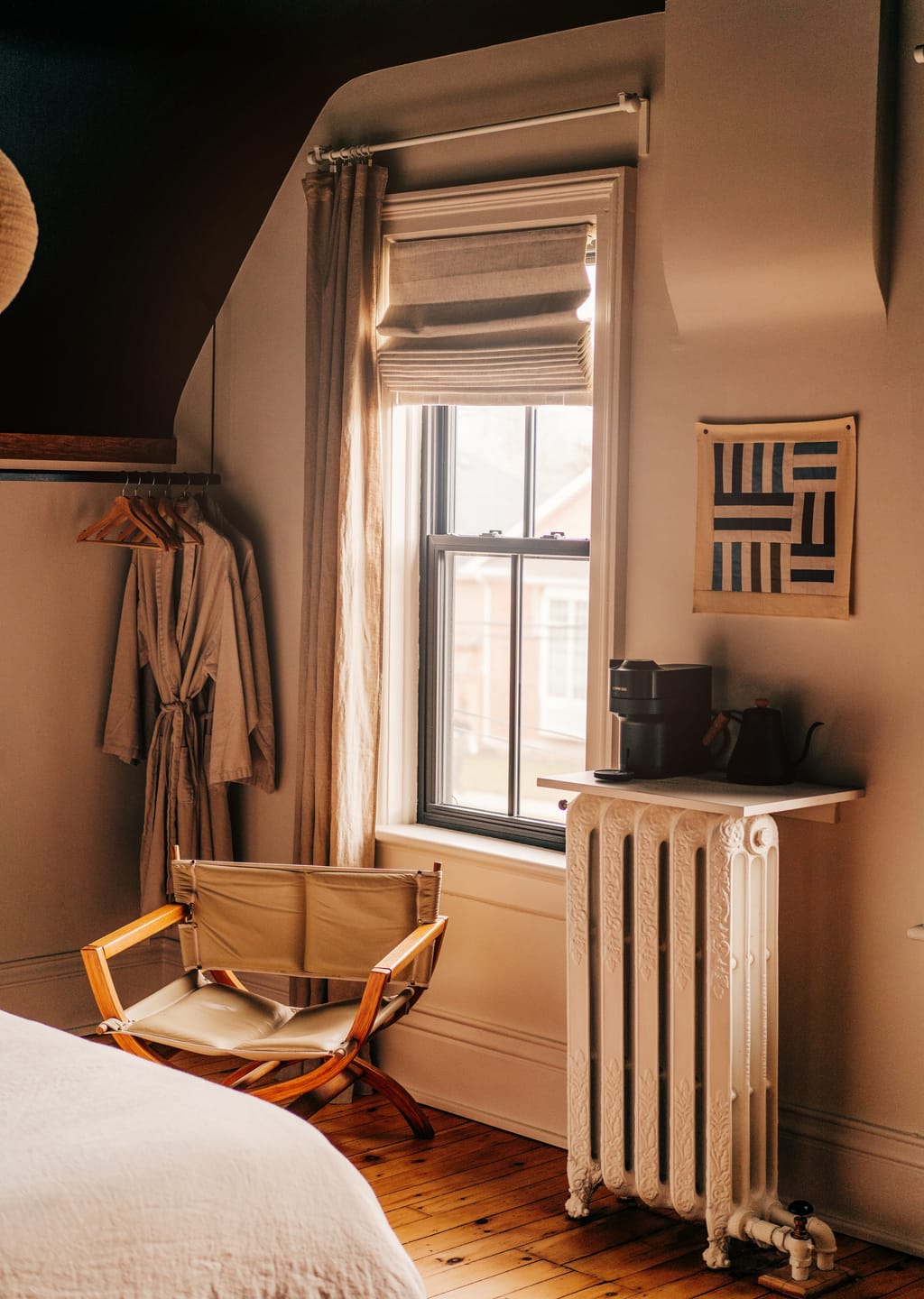 Guest room window with linen curtains, a folding leather chair, robes on a rail and a radiator coffee station