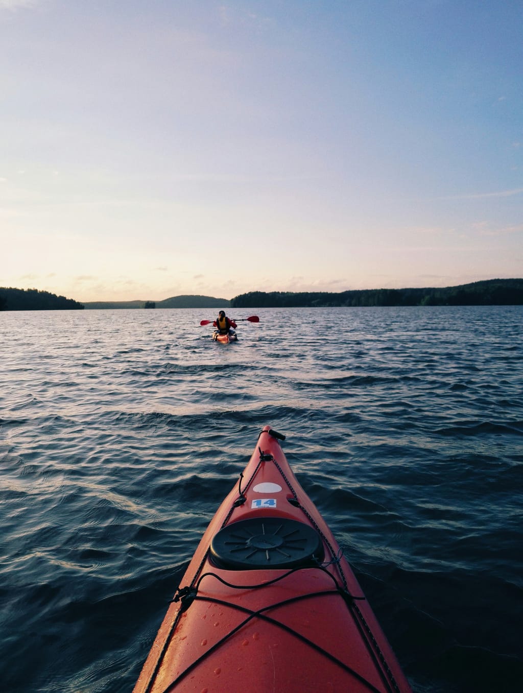 View from the bow of a red kayak on a calm lake at dusk, another paddler ahead