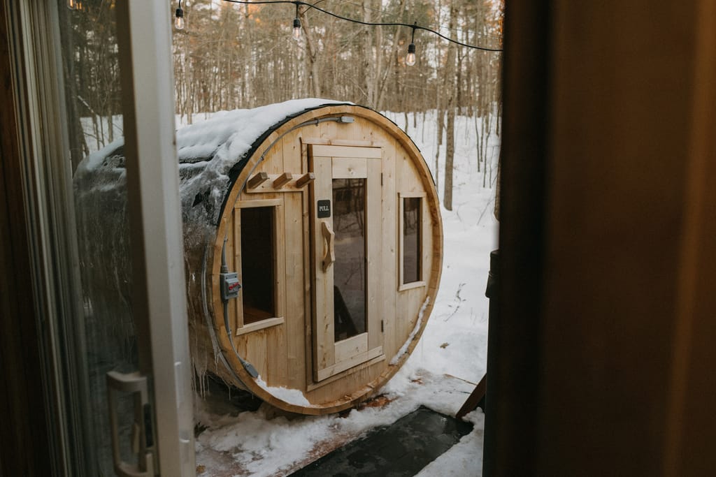 Barrel sauna in a snowy wood seen through an open glass door, string lights in the trees