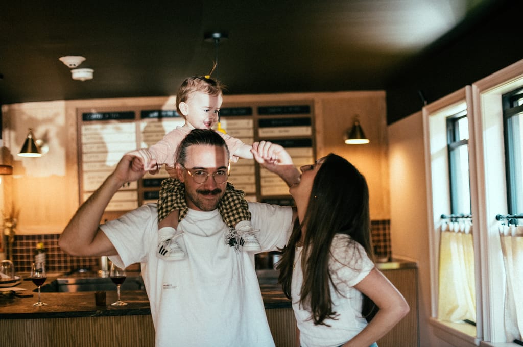 Toddler on a man's shoulders as a woman reaches up to hold hands, wine bar behind them