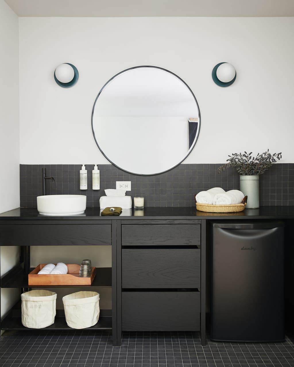 Black vanity with a round mirror, white vessel sink, towel basket and mini fridge