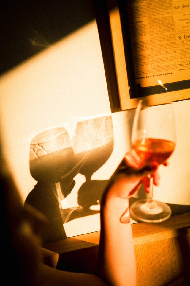 Hand raising a glass of orange wine, its shadow cast on a sunlit wall beside a framed vintage newspaper