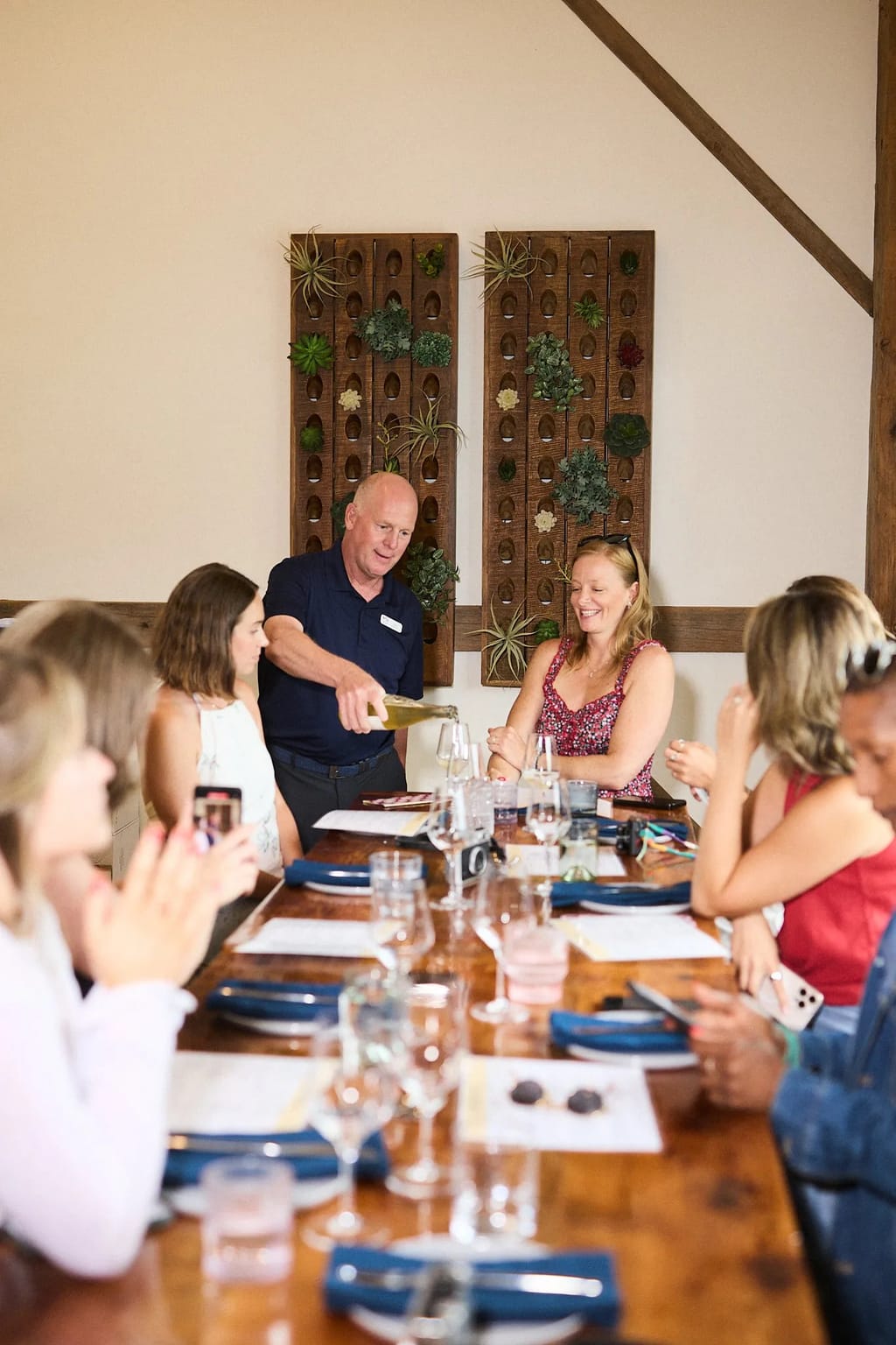 A host pours wine for guests seated along a long wood table at a hosted tasting