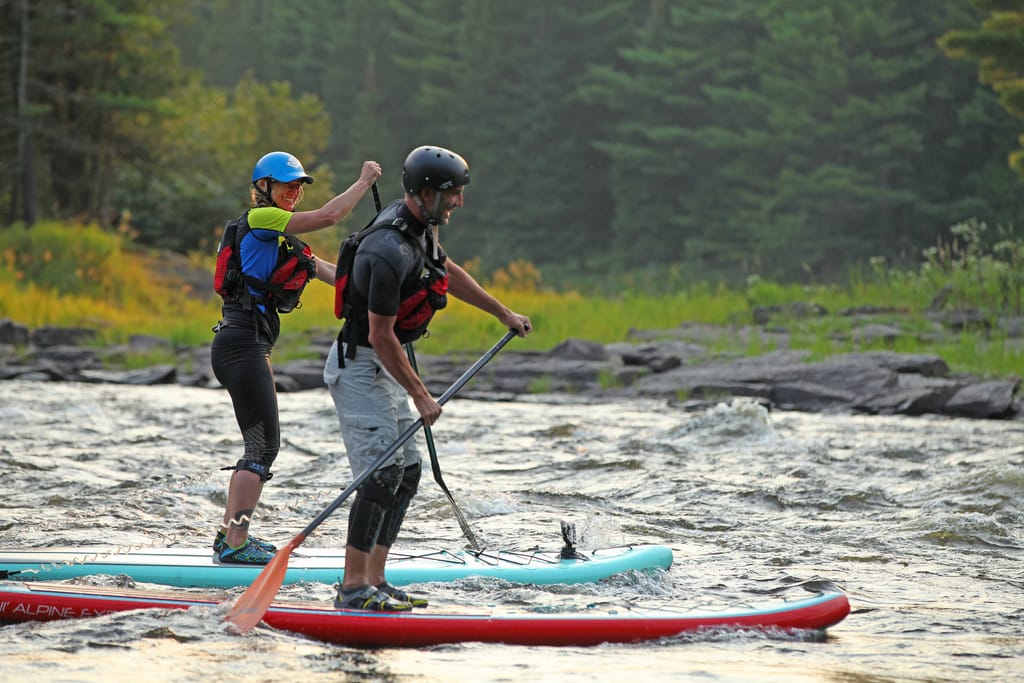 Two paddleboarders in helmets and life jackets standing on boards on a fast river, pines behind