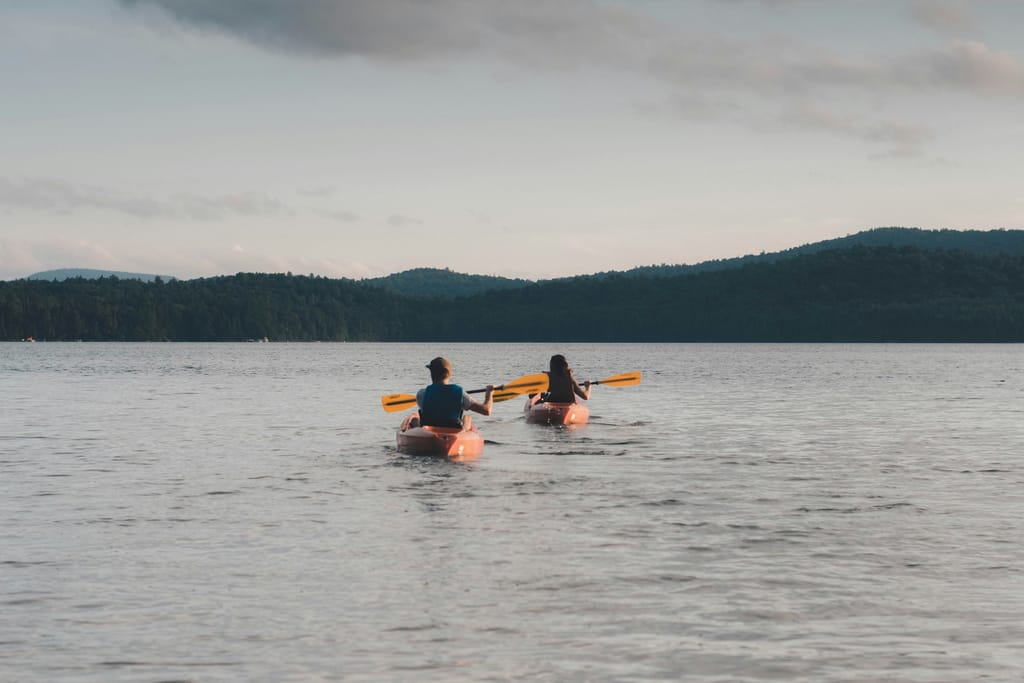 Two people paddling orange kayaks across a calm lake toward forested hills at dusk