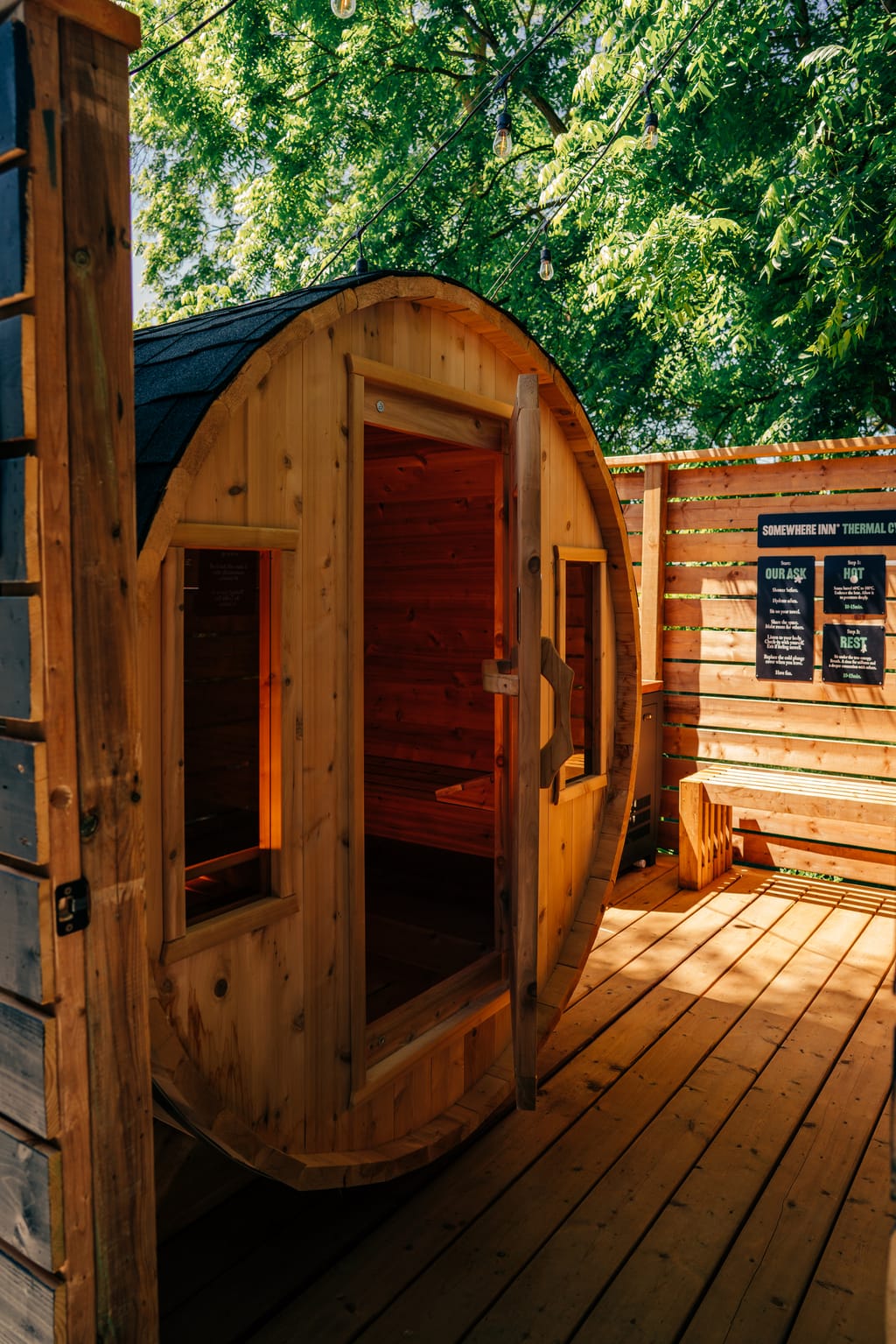 Barrel sauna on a wood deck with its door open, beside rules signs and a bench