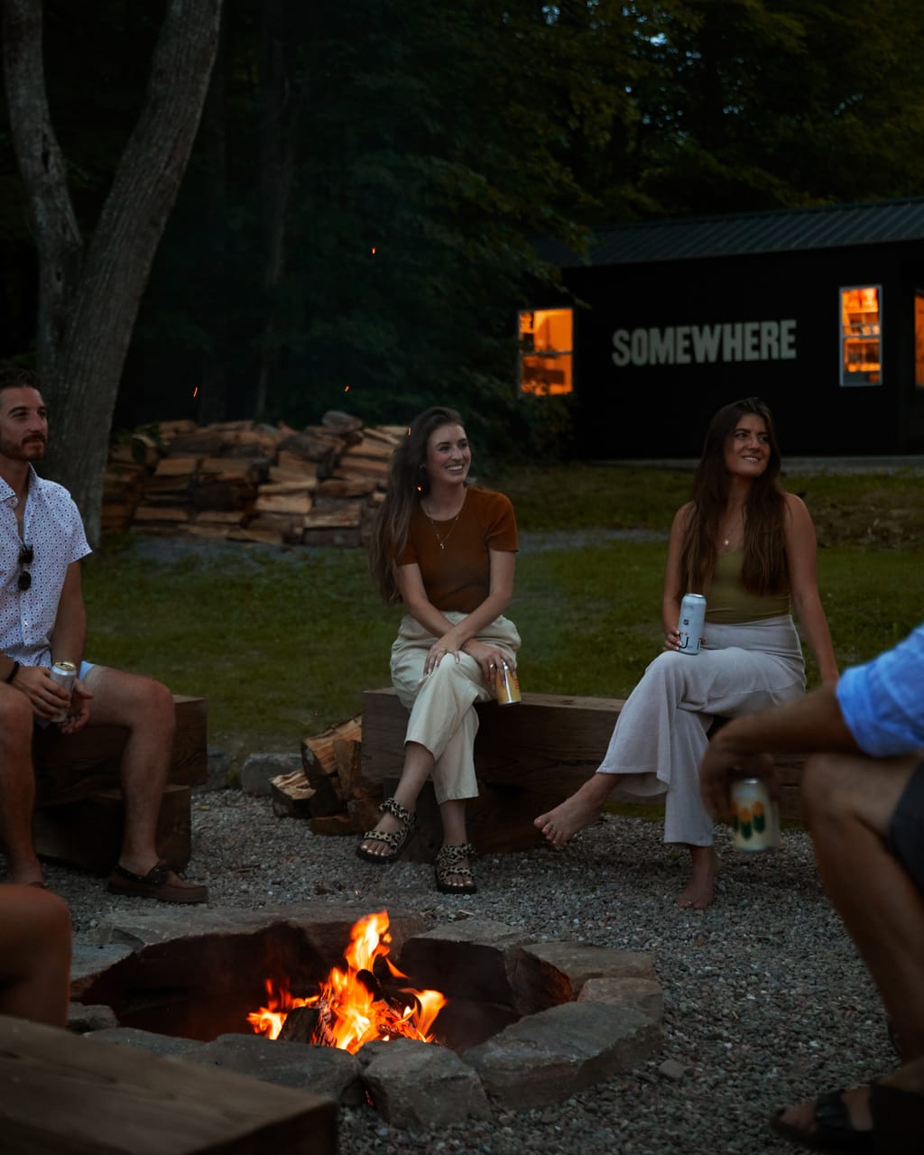 Guests sitting on timber benches around a fire pit at dusk outside a black cabin