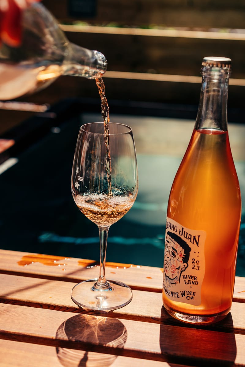 Orange wine poured into a glass beside the bottle, on a wooden ledge above dark water