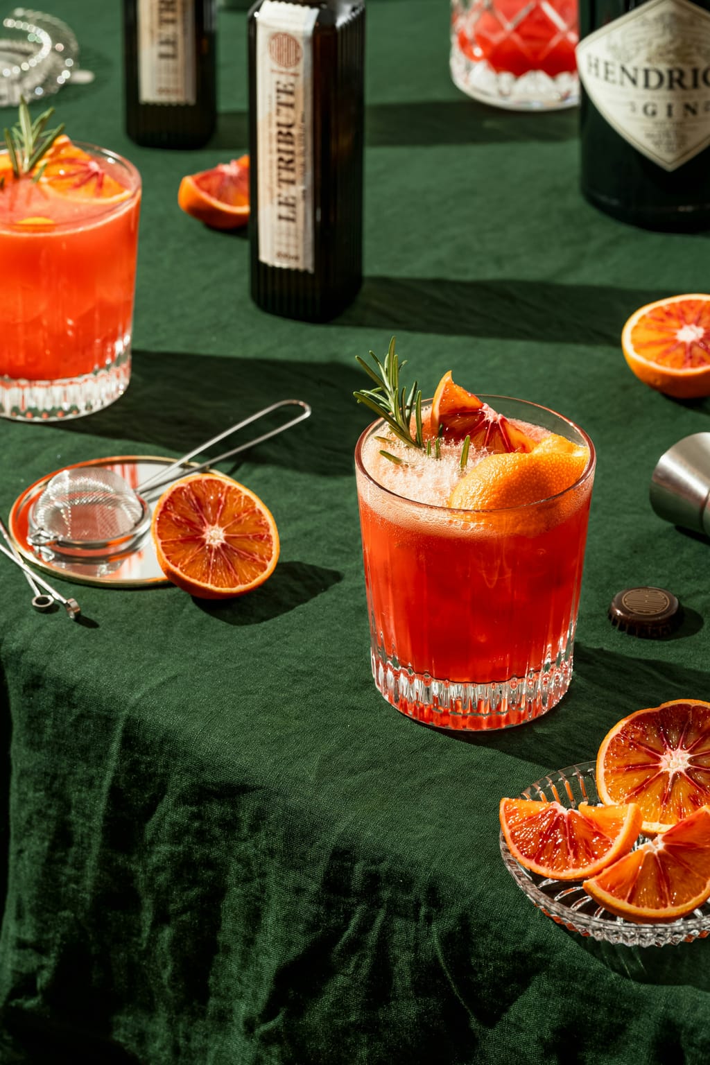 Blood orange cocktails with rosemary on a green linen table, bottles and citrus halves around them