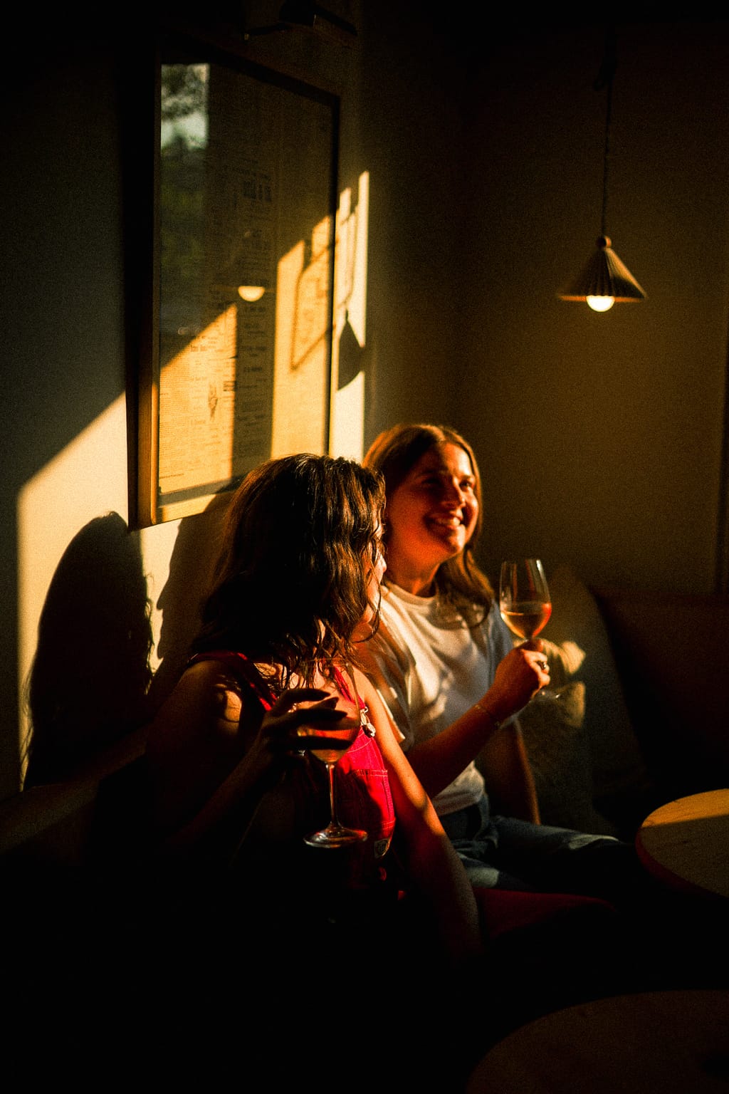 Two women smiling over glasses of wine on a banquette in a sunbeam under a pendant lamp