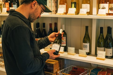 Man in a cap reading a wine bottle label beside a shelf of bottles in a shop