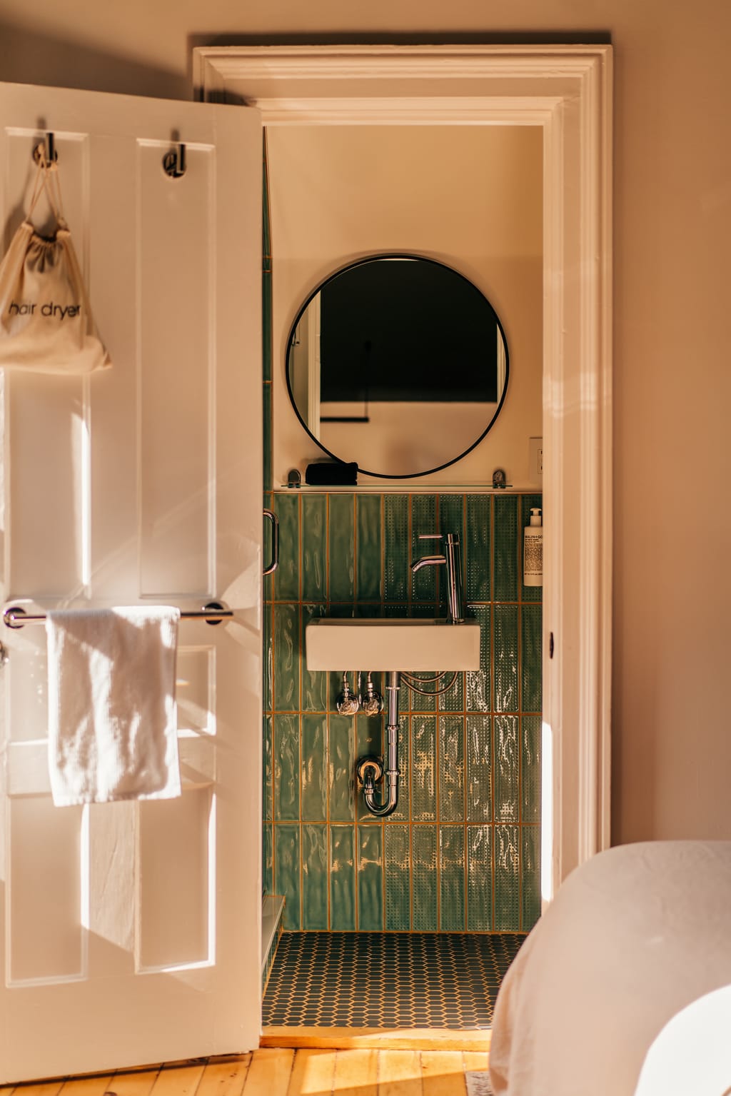 Small bathroom with green tile, a round mirror and a wall-hung sink seen through a door