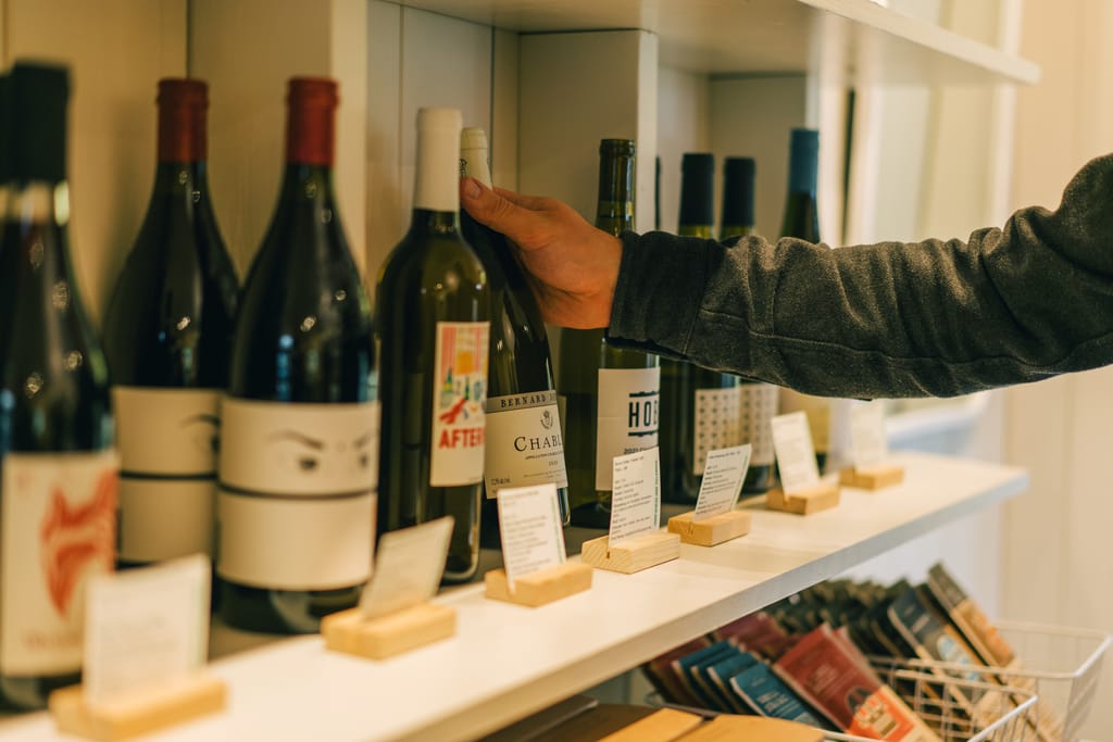 A hand reaches for a bottle on a white shelf of wines with small cards, chocolate bars below
