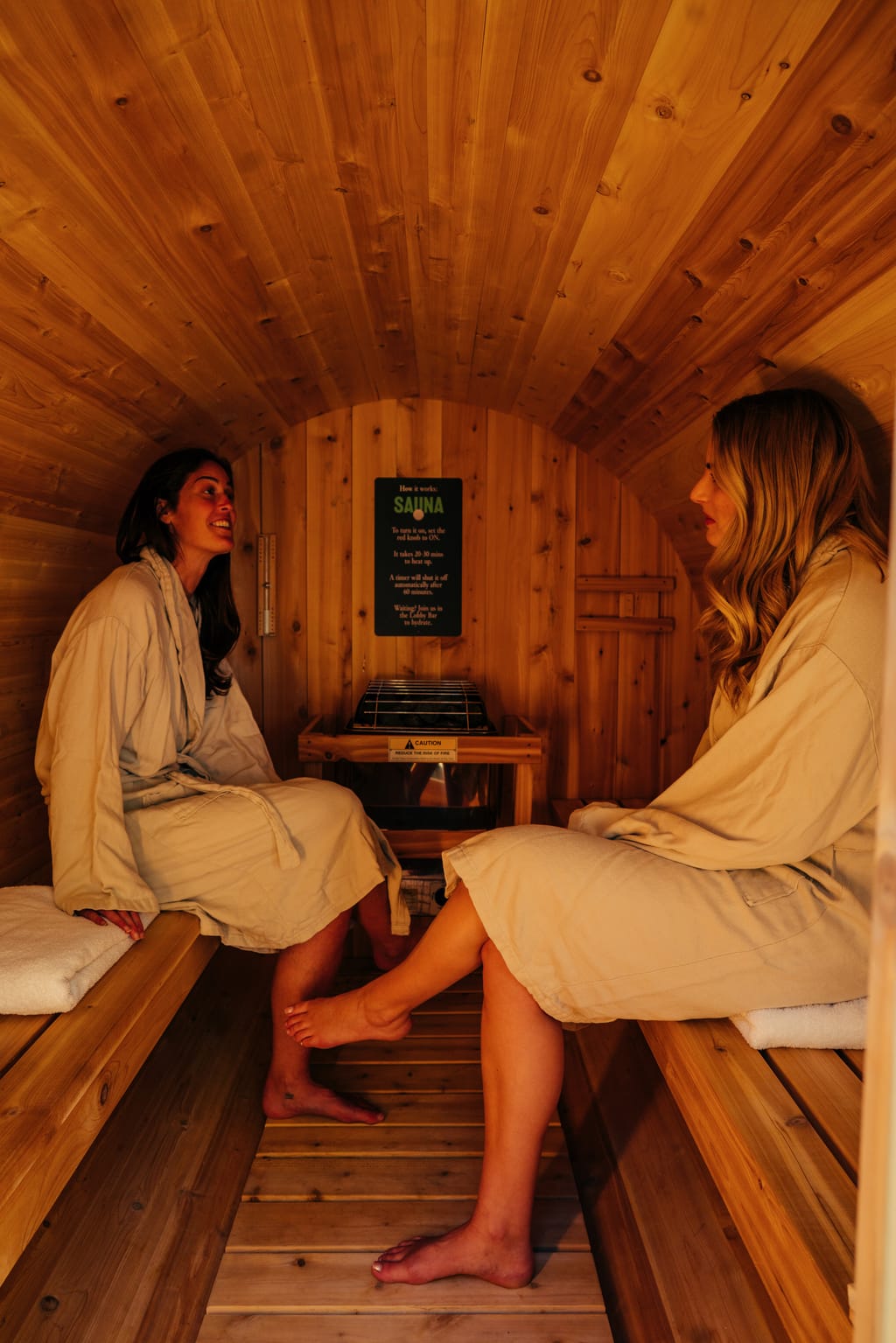 Two women in robes talking on the benches inside a cedar barrel sauna
