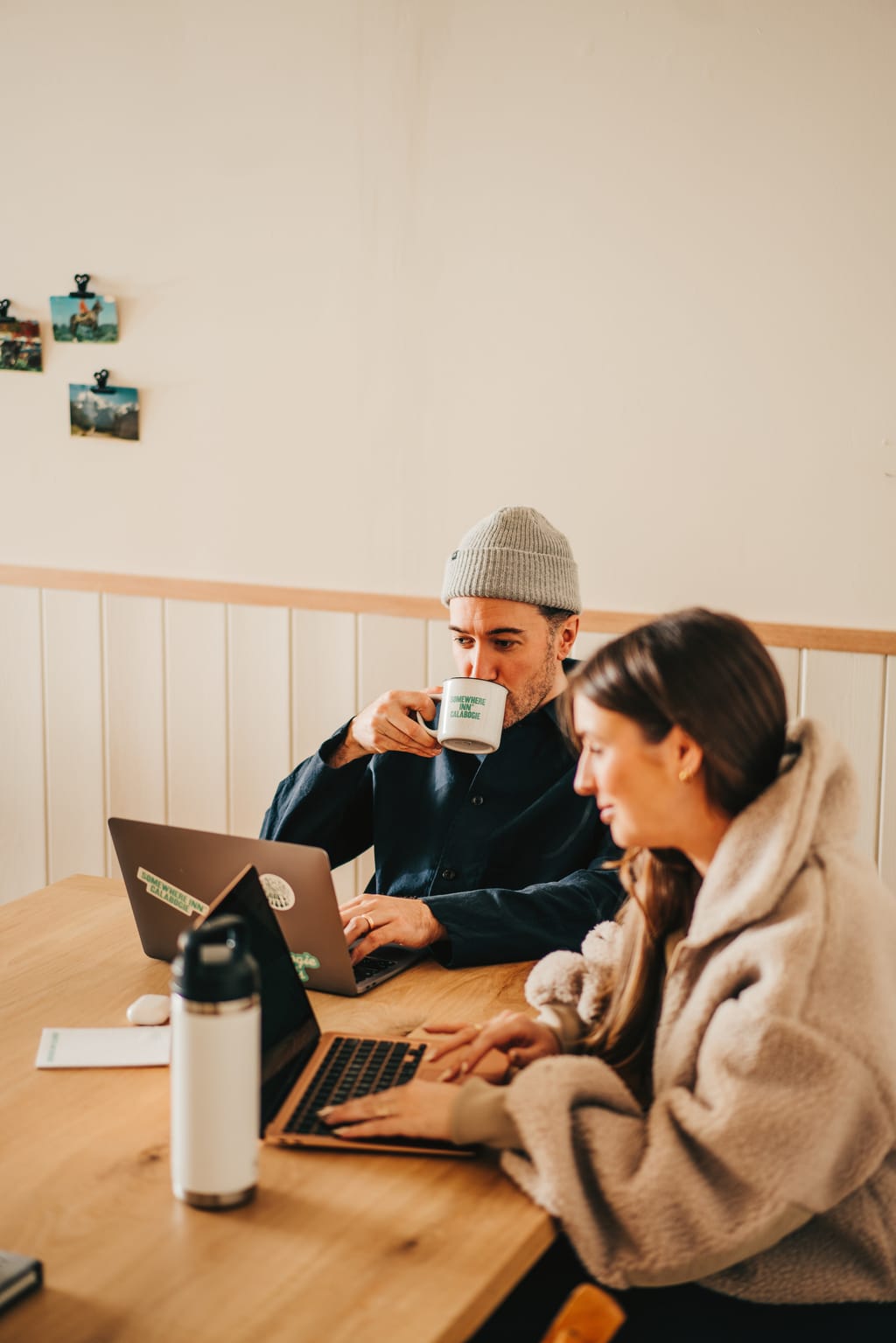 Two people work on laptops at a wood table, one sipping from a Somewhere Inn Calabogie mug