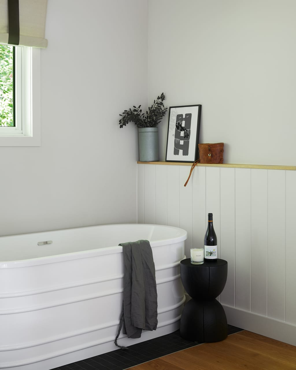 Freestanding white tub with a grey linen towel, a stool holding a bottle of wine and a candle