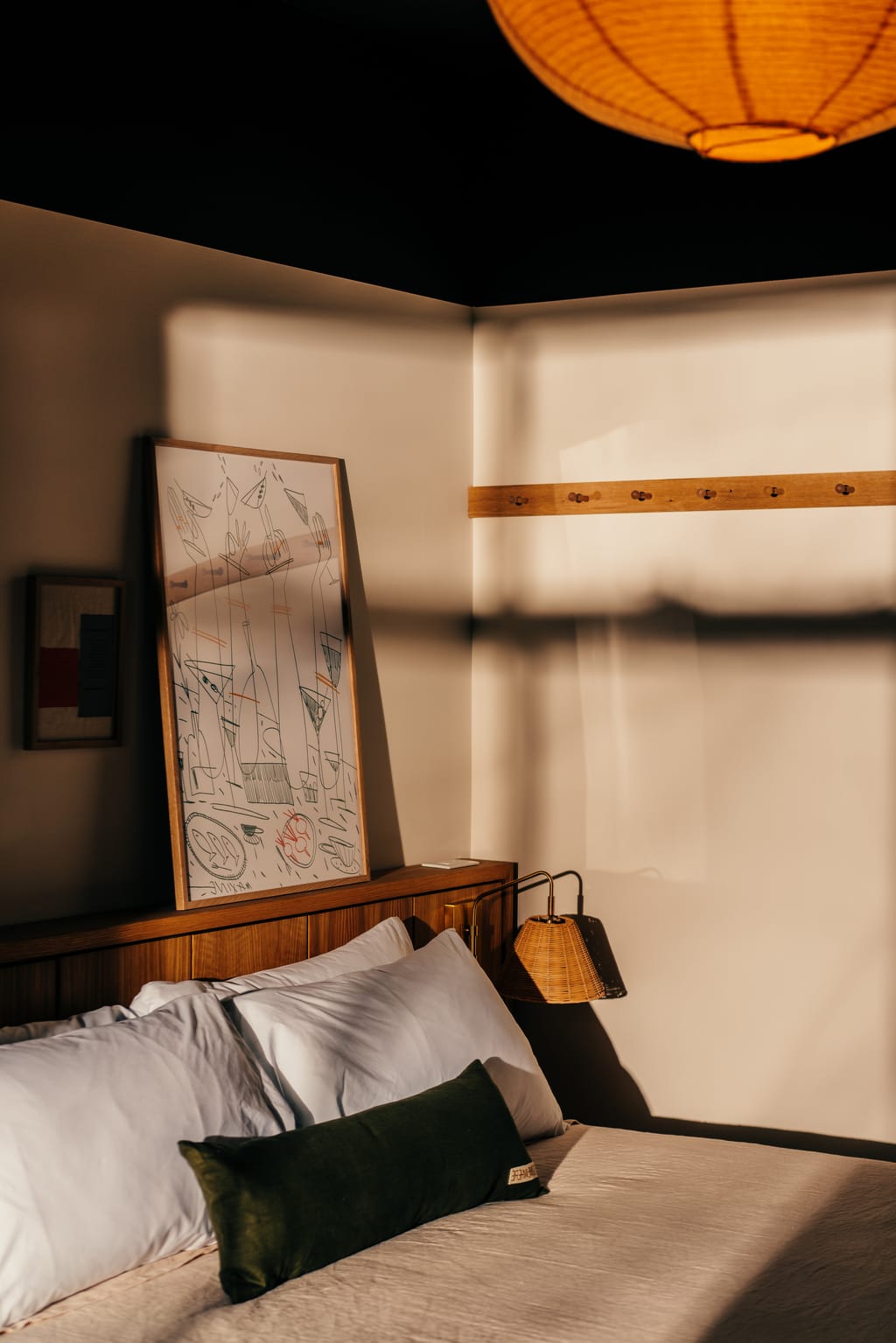 Bed corner with white pillows, a green bolster, framed drawing and a rattan wall lamp in window light