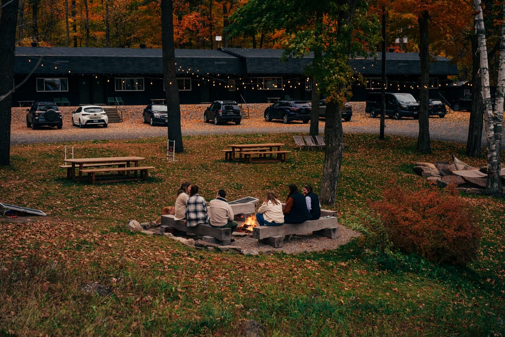 Friends around a fire pit in autumn at Somewhere Inn Calabogie, string lights over the black boutique motel behind