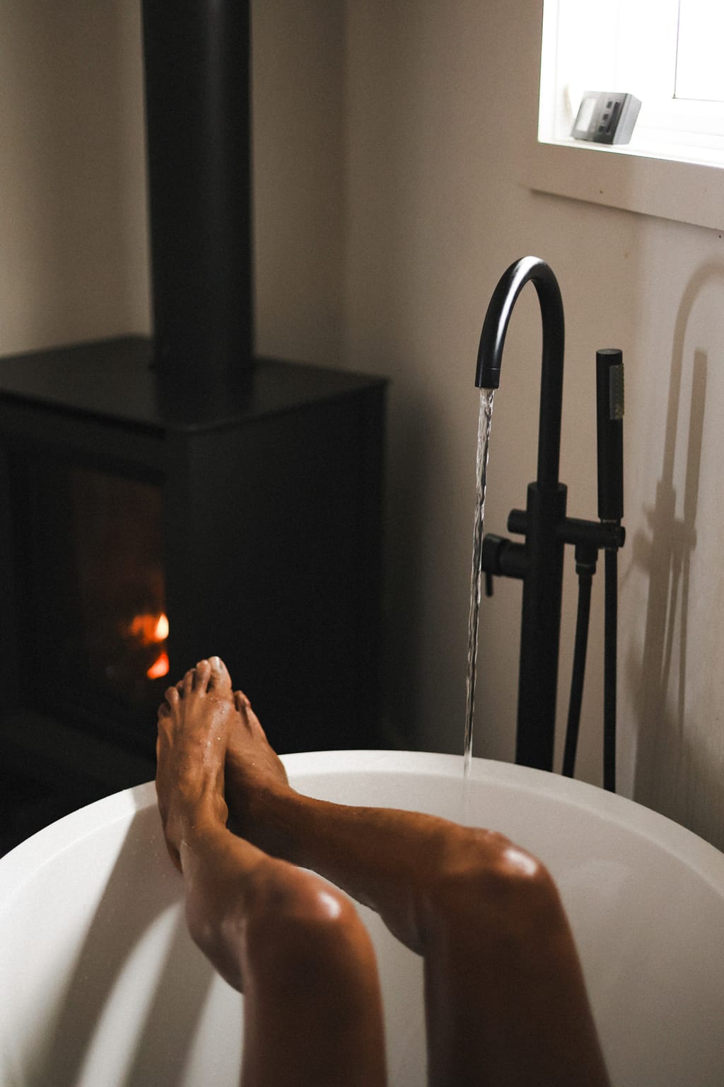 Crossed feet resting on the rim of a freestanding tub as the tap runs, a wood stove glowing behind