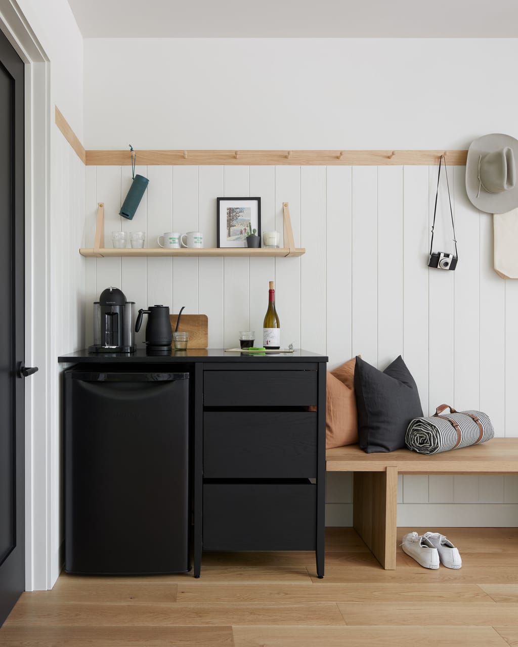 Room nook with a coffee station, mini fridge, wall pegs with a camera and hat, and a wood bench