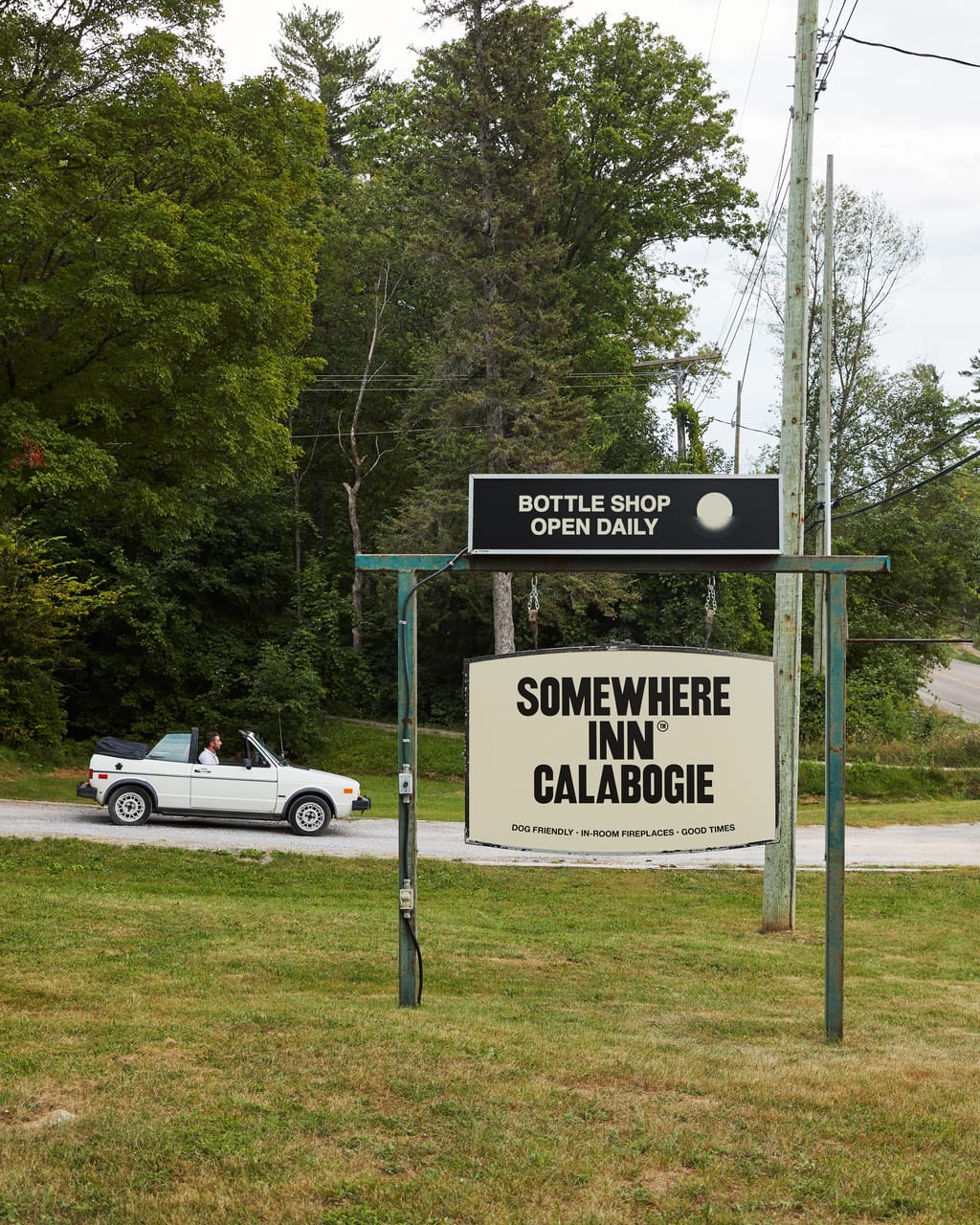 Somewhere Inn Calabogie roadside sign with a bottle shop banner and a white convertible passing