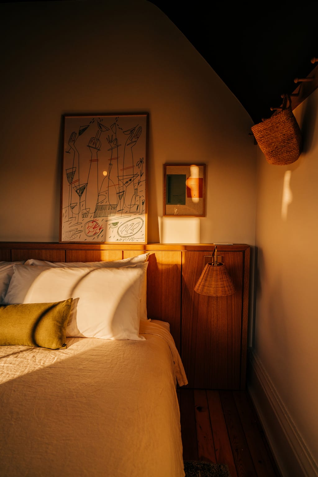 Bed with a wood headboard and a framed line drawing, lit by low golden sun