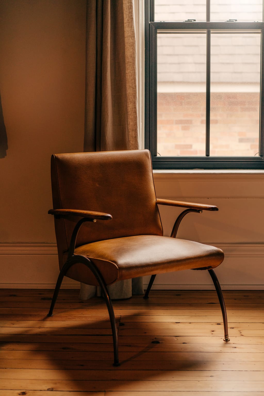 Leather armchair with wooden arms beside a window and linen curtain on a pine floor