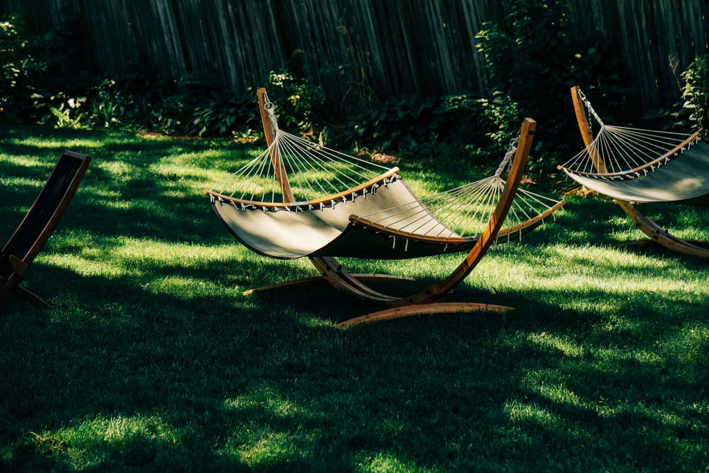 Two wooden-framed hammocks on a shaded green lawn beside a tall fence