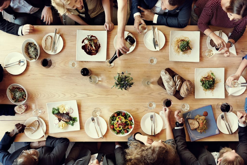 Overhead view of a long wooden table set with shared plates, wine glasses and many hands reaching in
