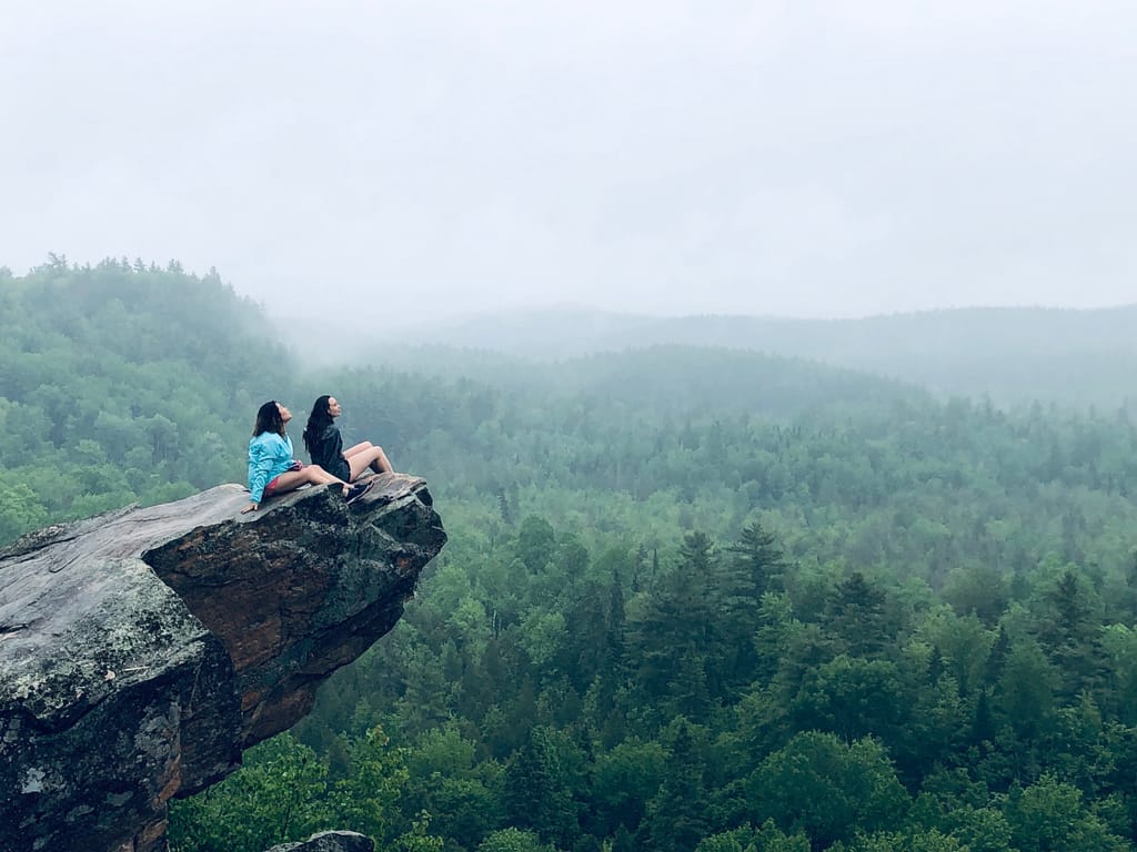 Two people sitting on a rock outcrop above a misty valley of forested hills