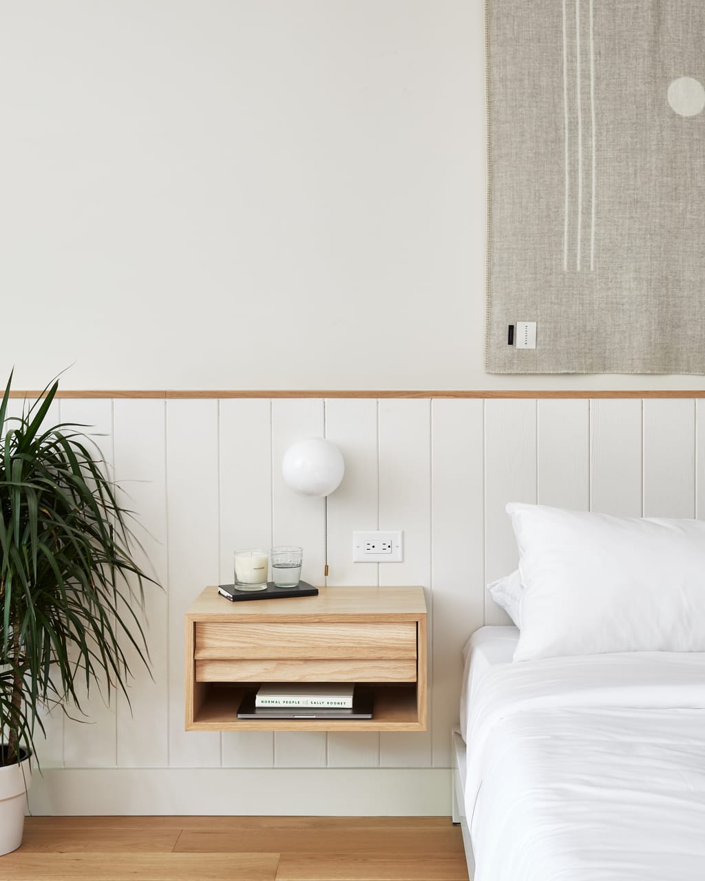 Wall-hung oak nightstand with a candle and book beside a bed, white panelling and a potted plant