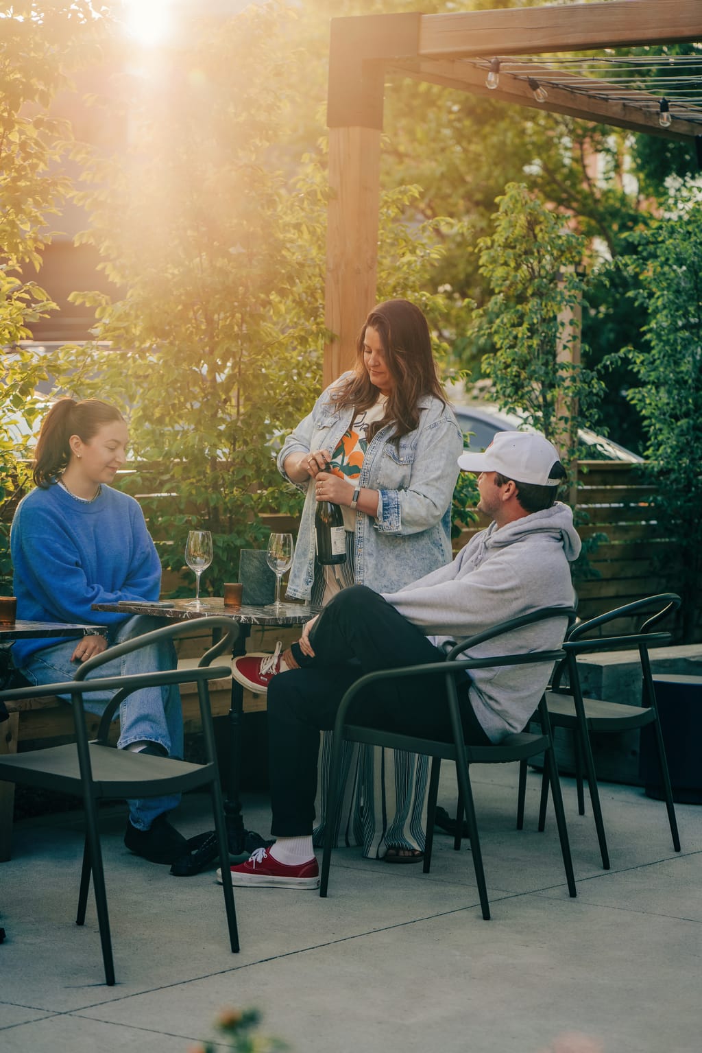 Woman opening a bottle of sparkling wine for two guests seated on a sunny patio