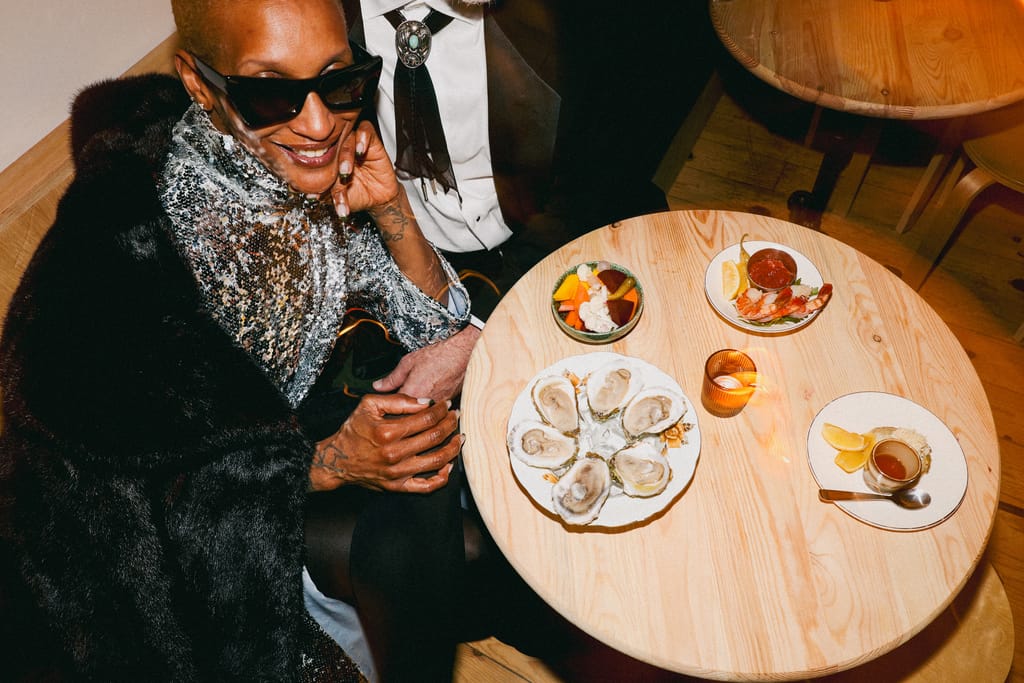 Woman in sequins and sunglasses at a round table set with oysters, shrimp and small plates