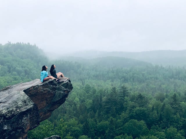 Two women sitting on a rock ledge looking out over misty forested hills